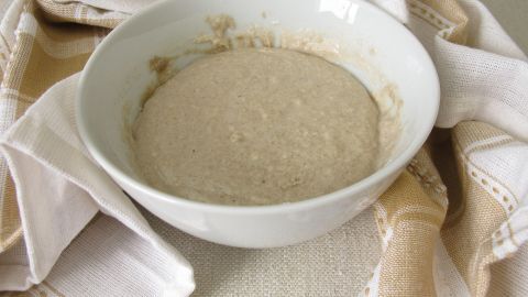 Ein Schüssel mit Teig steht auf einer Tischoberfläche und wird von einem Küchentuch bedeckt, um während der Ruhezeit besser zu gehen.