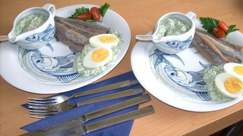 Zwei Teller mit frischer Matjes, gehackten Eiern und Grie Soß auf einem Holztisch, umgeben von Besteck und frischen Tomaten.