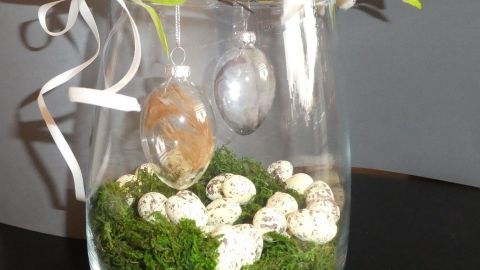 Ein Glasgefäß mit Moos und kleinen Eiern, dekoriert mit modernen Ostereiern und einem Zweig, der frisch wirkt.
