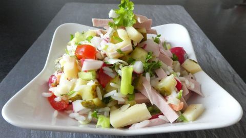 Ein bunter Wurstsalat wird auf einem weißen Teller präsentiert, umgeben von frischen Gemüsezutaten und Kräutern.