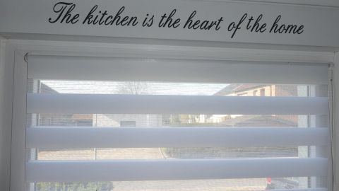 Ein Doppelrollo ist halb hochgezogen und lässt Licht in eine helle, moderne Küche, während darüber der Spruch "The kitchen is the heart of the home" steht.