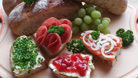 Vier knusprige Brötchen liegen auf einem Teller, umgeben von bunten belegten Broten und Trauben.