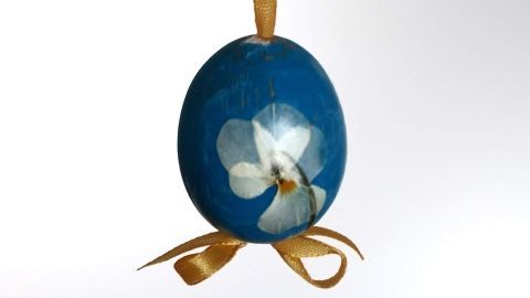 Ein blaues Osterei mit einer Blüte hängt dekorativ an einem gelben Band von einem Zweig, vor hellem Hintergrund.