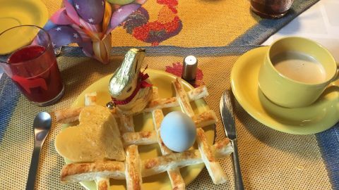 Ein gebackenes Osternest aus Blätterteig mit einem Ei-Herz liegt auf einem dekorierten Tisch, umgeben von Getränken und Geschirr.