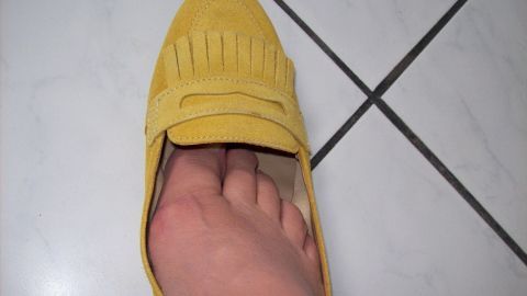 Ein gelber Schuh liegt auf einem Fliesenboden, während ein Fuß mit sichtbaren Blasen darin steckt, was Unbehagen zeigt.