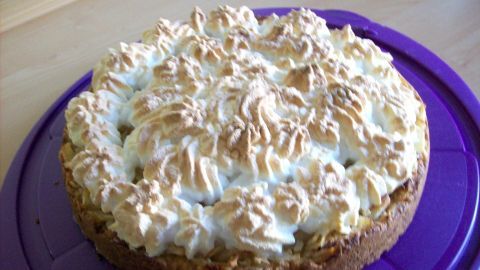 Eine Apfeltorte mit einer goldbraunen Baiserhaube liegt auf einem lila Teller, umgeben von einer neutralen Küchenoberfläche.