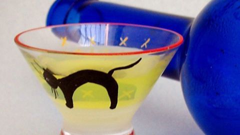 Ein bunter Likör in einem Glas mit Katzendesign steht neben einer blauen Flasche auf einem neutralen Hintergrund.