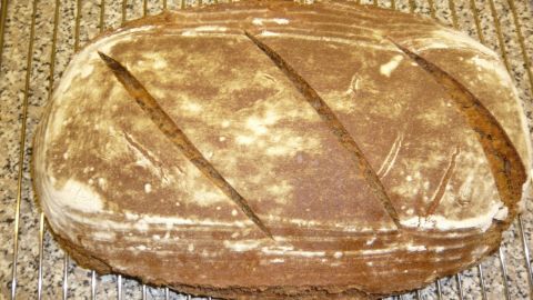 Ein frisch gebackenes Sauerteig-Roggenmischbrot mit einer goldbraunen Kruste liegt auf einem Kuchengitter in einer hellen Küche.