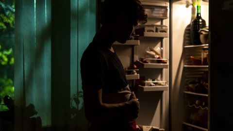 Eine Person steht nachtblind vor einem offenen Kühlschrank und betrachtet den Inhalt, umgeben von schwachem Licht.
