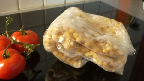 Eine durchsichtige Verpackung mit Hülsenfrüchten liegt auf einem dunklen Kochfeld neben frischen Tomaten in einer Küche.