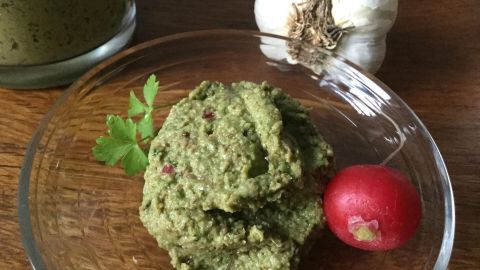Ein grüner, cremiger Pesto-Haufen aus Radieschen und ihrem Grün liegt in einer Schale, umgeben von einer Knoblauchzehe und einer Radieschen.