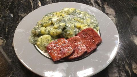 Auf einem grauen Teller liegen Dillkartoffeln, umgeben von einer cremigen Soße, und einige Scheiben Fleisch.