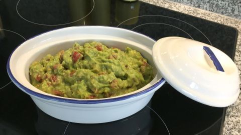 Ein gefüllter, weißer Behälter mit einem Avocado-Tomaten-Dip steht auf einer Herdplatte aus schwarze. Der Deckel ist teilweise geöffnet.