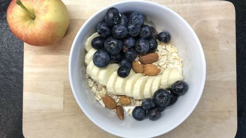 Eine Schüssel mit Haferflocken, Banane, Heidelbeeren und Mandeln steht auf einem Holzbrett, neben einem Apfel für Meal-Prep.
