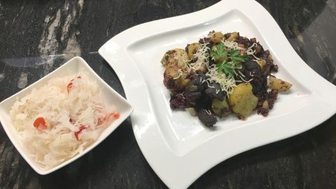 Ein herzhaftes Blunzengröstl auf einem eleganten Teller, umgeben von einem Schälchen mit Salat, auf einem dunklen Tisch.