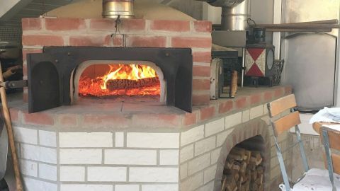 Ein Lehmbackofen mit loderndem Feuer backt Pizza in einem rustikalen Raum, umgeben von Holzvorräten und viel Licht.