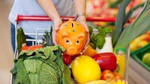 Eine Hand hält ein Sparschwein über einem vollen Einkaufswagen mit frischem Gemüse und Obst im Supermarkt.