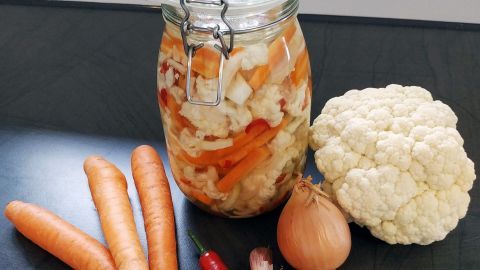Ein Glas mit fermentiertem Gemüse steht neben frischen Karotten, Blumenkohl, Zwiebel und einer Chilischote auf einer dunklen Oberfläche.