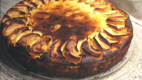 Eine Apfel-Käse-Torte ohne Boden liegt auf einem Teller und ist mit goldbraunen Apfelscheiben dekoriert.