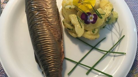 Geräucherte Makrele liegt auf einem Teller neben einem Kartoffelsalat, dekoriert mit Kräutern und Blüten.