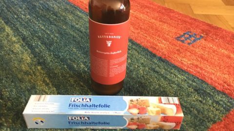 Eine braune Flasche mit einer roten Etikette steht auf einem bunten Teppich neben einer Packung mit Frischhaltefolie.