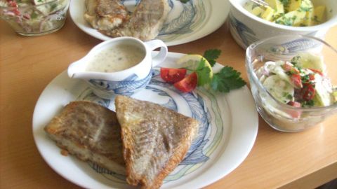 Gebratener Seelachs liegt auf Tellern, umgeben von Bechamel-Soße und frischem Gemüse in Schüsseln auf einem Tisch.