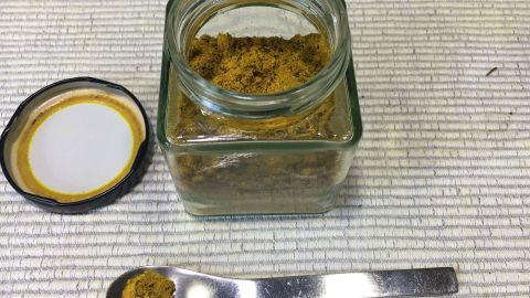 Ein Glasbehälter mit Kurkuma steht offen auf einer Tischoberfläche, während ein Löffel Kurkuma daneben platziert ist.