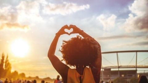 Eine Person mit lockigem Haar formt ein Herz mit den Händen, während sie vor einer Menge bei Sonnenuntergang steht.