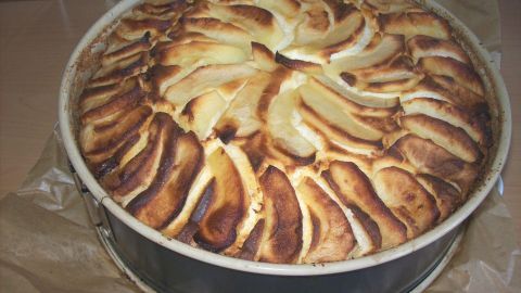 Ein Käsekuchen mit goldbraunen Rum-Apfelschnitzen, frisch gebacken, steht auf Backpapier in einer runden Springform.