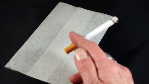 Eine Hand hält eine Zigarette über einem Lenor-Tuch, um Gerüche im Wohnraum zu beseitigen. Der Hintergrund ist schwarz.