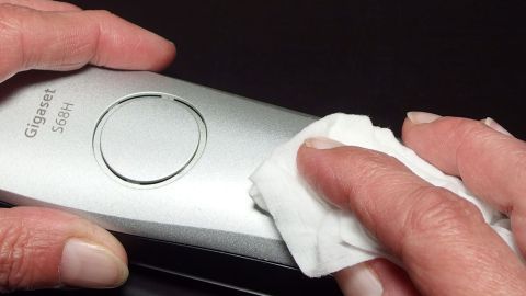 TV, Telefon, Fernbedienung etc. - Staubwischen leicht gemacht. Eine Hand wischt mit einem Tuch über eine Fernbedienung, um Staub zu entfernen, umgaben von einem sauberen Tisch.