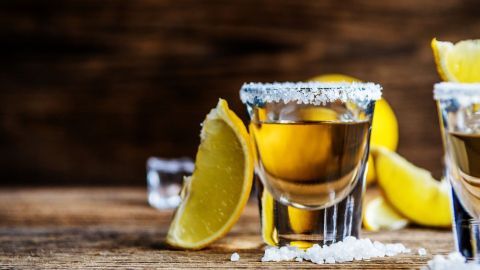 Gläser mit Tequila stehen auf einem Holztisch, umgeben von Limettenscheiben und Salz, bereit zum Servieren.
