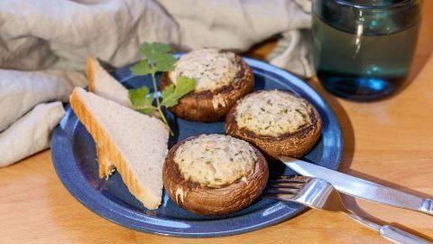 Gefüllte Champignons liegen auf einem blauen Teller neben Brotscheiben, umgeben von einem Glas Wasser und einem Tuch.