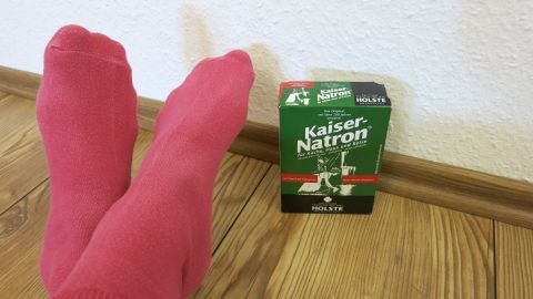 Rosa Socken liegen entspannt auf einem Holzboden neben einer grünen Natronverpackung, um Schweißfüße zu behandeln.