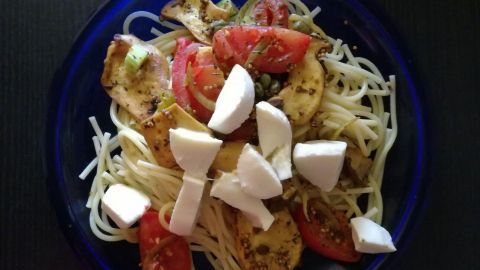 Eine bunte Schüssel Spaghetti mit Kräuterseitlingen, frischen Tomaten und Mozzarella auf einem dunklen Tisch.