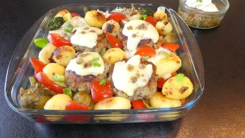 Käseüberbackene Pesto-Frikadellen liegen auf einem bunten Gemüsebett aus Kartoffeln, Tomaten und Brokkoli in einer Glasschale.
