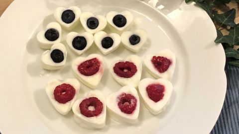 Herzförmige gefrorene Joghurtherzen mit Himbeeren und Heidelbeeren liegen auf einem Teller, umgeben von grünem Blattwerk.