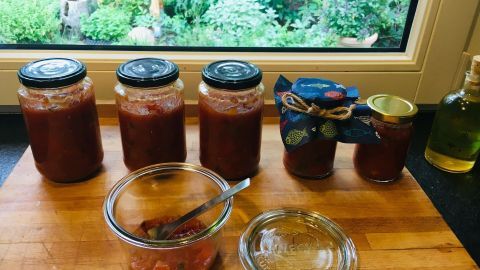 Gläser mit scharfem Tomaten-Paprika-Chutney stehen auf einem Holzbrett, umgeben von einer grünen Gartenlandschaft.