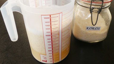 Ein Messbecher mit einer Mischung aus Kokosmilch steht neben einem Glas mit Kokosmehl auf einem Tisch in einer Küche.