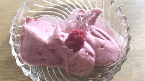 Eine Schüssel mit cremiger, rosa Nicecream aus Bananen, garniert mit einer Himbeere, auf einem Holztisch.