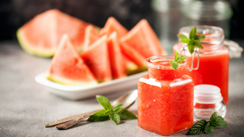 Ein Glas Wassermelonenmarmelade steht vorne, während frisch geschnittene Melonenscheiben im Hintergrund liegen und Minzblätter dekorieren.