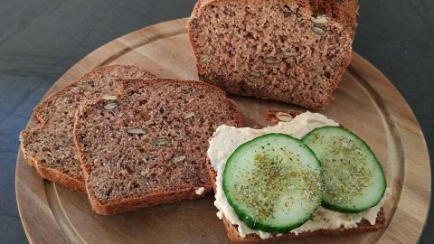 Ein frisch gebackenes Vollkornbrot liegt auf einem Holzbrett. Eine Scheibe ist mit Frischkäse und Gurkenscheiben belegt.