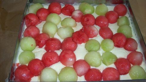 Ein saftiger Kuchen mit Melonenstückchen liegt in einem rechteckigen Behälter, umgeben von einer hellen Sahneschicht.