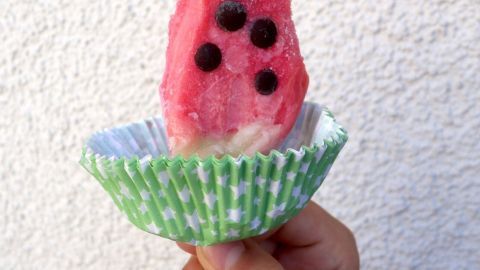 Ein Hand hält ein Eis in Form einer Wassermelone, das in einer grünen Waffelschale steht, vor einer weißen Wand.