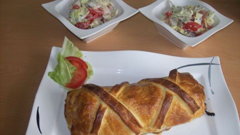 Ein gebackenes Schweinefilet im knusprigen Blätterteig liegt auf einem Teller, umgeben von zwei Schalen mit Salat.