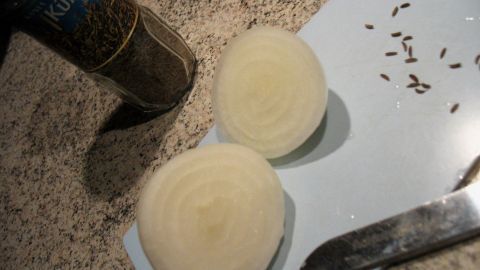 Eine halbierte Zwiebel liegt auf einem Schneidebrett, während ein Messer daneben und Gewürze im Hintergrund sichtbar sind.