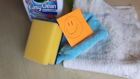 Eine gelbe Schwamm und ein blauer Reinigungstuch liegen neben einem Reinigungsmittel und einem Post-it mit einem Smiley.