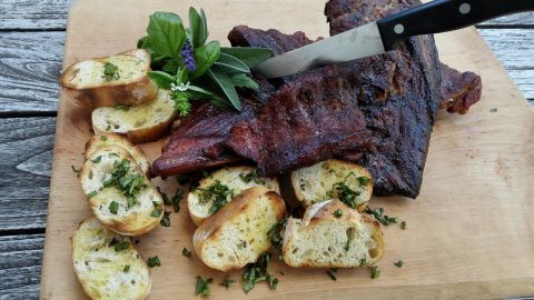 Auf einem Holzbrett liegen saftige Spareribs, während ein Messer sie anschneidet, umgeben von knusprigem Brot und Kräutern.
