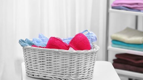Rosa und blaue BHs liegen ordentlich in einem weißen Korb auf einem Tisch, umgeben von einem hellen Raum mit Wäsche.