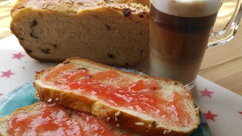 Ein fluffiger Hefezopf liegt hinter zwei Scheiben Brot mit Marmelade, begleitet von einem Glas Kaffee auf einem hellen Tisch.
